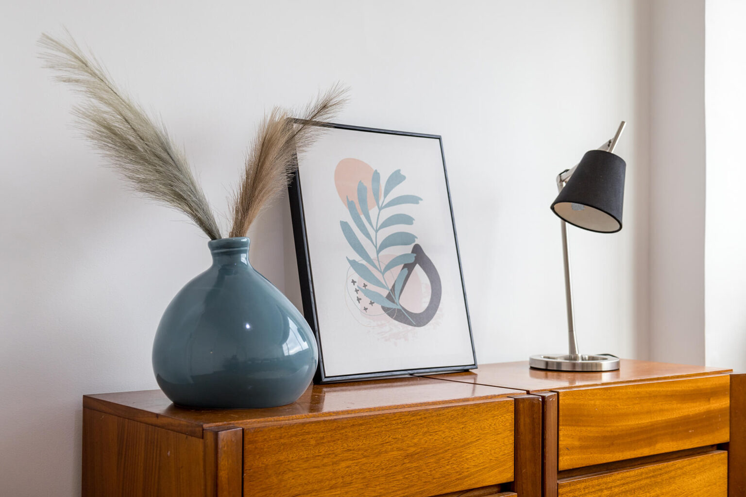 Arranjo decorativo em cima de um móvel de madeira com vaso azul, ilustração moderna e candeeiro elegante.