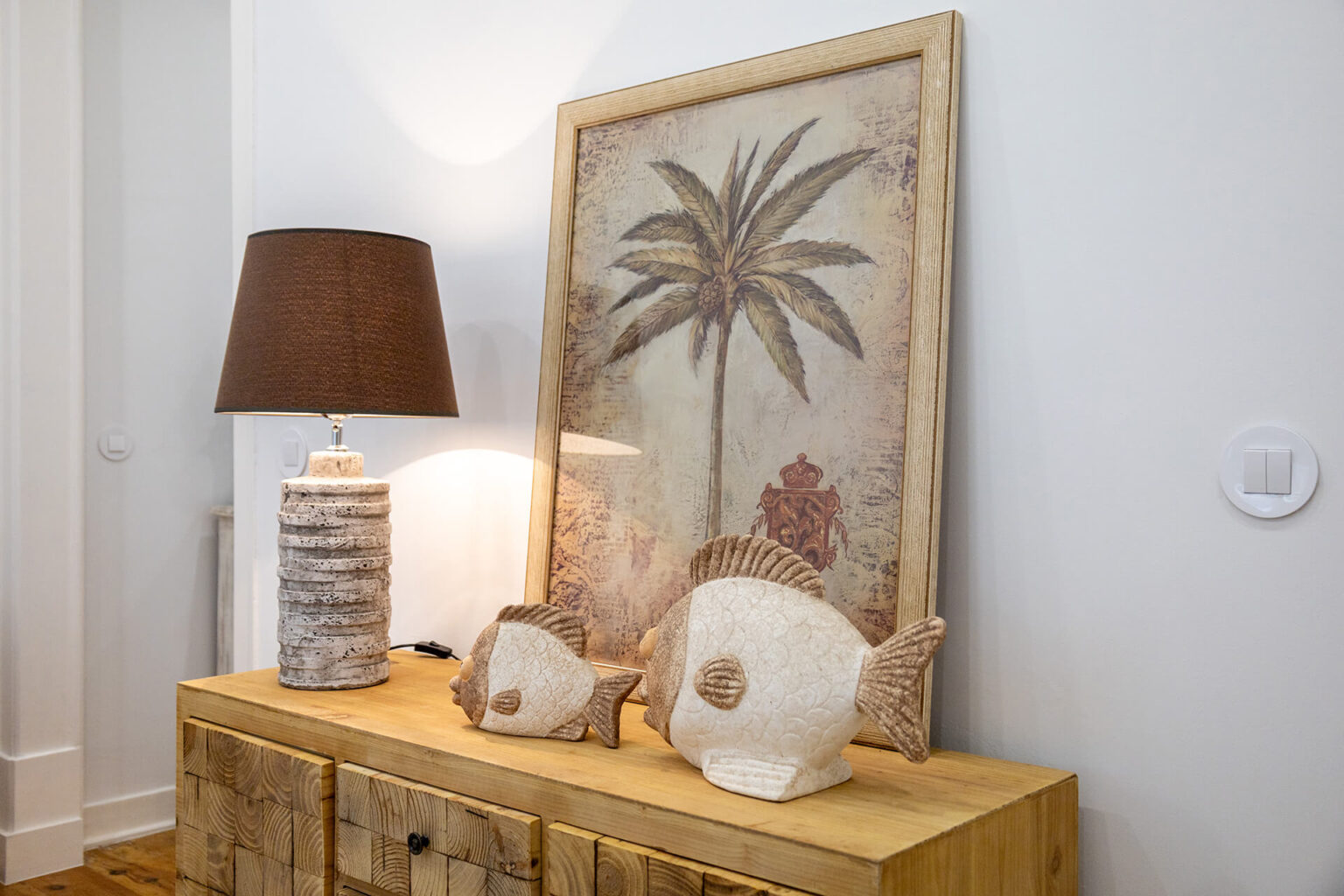 Decoração de aparador em estilo costeiro com uma lâmpada texturizada, quadro de palmeira e peças decorativas de peixe.